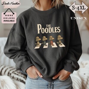 Può includere: Felpa unisex grigio scuro con la scritta "The Poodles" e un'illustrazione di quattro barboncini che attraversano le strisce pedonali. La felpa ha la scritta "S-4XL" e "Unisex" in bianco. È presente anche la scritta "More Colors Available".