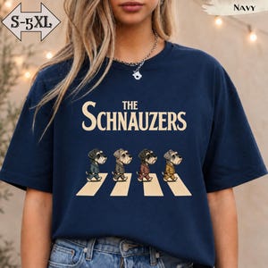 May include: A navy blue t-shirt featuring the text THE SCHNAUZERS in cream lettering above a graphic of four Schnauzer dogs walking across a crosswalk. The dogs are wearing suits in shades of grey, brown, and yellow. The shirt is available in sizes S-5XL.