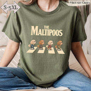 Peut inclure: Un t-shirt vert olive avec le texte "THE MALTIPOOS" en police couleur crème au-dessus d'une illustration de quatre chiens Maltipoo marron marchant sur des bandes de passage piéton blanches. L'étiquette indique les tailles S-5XL.