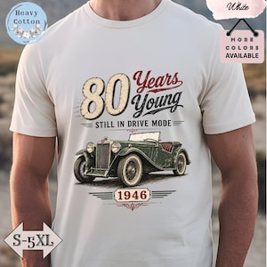 Op de afbeelding: Crèmekleurig T-shirt met een vintage auto-illustratie en de tekst "80 Years Young Still in Drive Mode 1946." Het ontwerp bevat een klassieke auto in groen en rood, met het getal 80 in een groot lettertype.