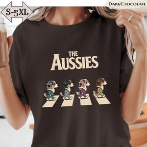 Peut inclure: Un t-shirt marron chocolat foncé avec le texte THE AUSSIES en lettres crème au-dessus d'un graphique représentant quatre chiens bergers australiens marchant sur un passage piéton, chacun portant une veste de costume, tailles S-5XL.