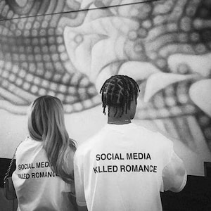 Könnte beinhalten: Schwarz-Weiß-Foto von zwei Personen, die weiße T-Shirts tragen. Der Text "SOCIAL MEDIA KLLED ROMANCE" ist in Schwarz aufgedruckt. Im Hintergrund ist ein verschwommenes Wandbild zu sehen.