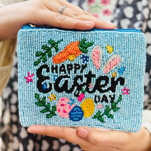 Könnte beinhalten: Hellblaue, perlenbesetzte Clutch mit Reißverschluss. Sie zeigt ein farbenfrohes Ostermotiv mit den Worten "HAPPY EASTER DAY" in schwarzen Perlen, sowie eine Karotte, Hasenohren und Ostereier.