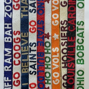Puede incluir: Correas de cuentas de varios colores, cada una con nombres de equipos y frases como "Go Saints" y "Go Vols". Las correas tienen cierres dorados y están diseñadas para bolsos o accesorios.