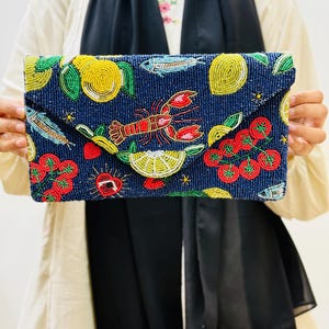 Puede incluir: Clutch azul marino con cuentas que presenta un diseño colorido de limones, tomates, peces y una langosta. El clutch rectangular tiene un cierre de solapa. Los adornos de cuentas añaden textura e interés visual al accesorio.