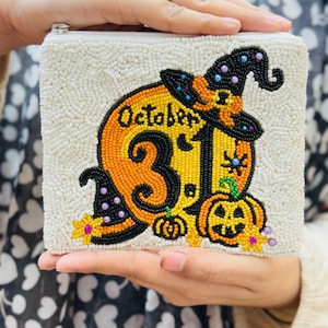 Op de afbeelding: Een witte, met kralen bezette clutch met een Halloween-ontwerp. Het ontwerp toont een pompoen met een heksenhoed, de tekst "October 31" en kleine pompoenen. De clutch wordt door een persoon vastgehouden.