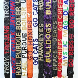 May include: A collection of beaded straps in various colors, each featuring team names and phrases like "TROY TROJANS" and "GO CATS" in gold or white lettering. The straps have gold-colored clasps.