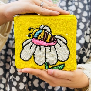 Peut inclure: Une pochette carrée jaune à sequins avec un motif d'abeille et de fleur en perles. L'abeille a des accents bleus, marron et roses. La fleur est blanche avec un contour noir et des feuilles vertes. La pochette a une fermeture éclair dorée.