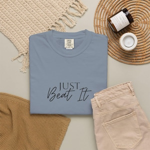 Op de afbeelding: Een lichtblauw t-shirt met de tekst "Just Beat It" in zwart script. Het shirt wordt getoond met een beige broek, een geweven placemat, een kaars en een decoratieve geweven mat.