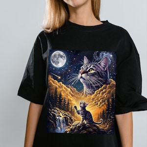 May include: A black t-shirt featuring a graphic print of a large tabby cat head in a starry night sky above a smaller tabby cat sitting on a rocky cliff. The scene includes golden clouds, a waterfall, a full moon, and a shooting star.