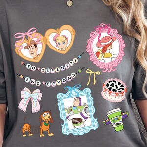 May include: Dark gray t-shirt featuring colorful cartoon character graphics and the text "TO INFINITY AND BEYOND". The design includes framed images of characters, a bow, a cake, and a coffee cup. The shirt is worn by a person.