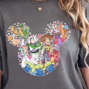 Puede incluir: Camiseta gris oscuro con un gráfico colorido de los personajes de Toy Story de Disney, dispuestos en forma de cabeza de Mickey Mouse. El diseño incluye a Buzz Lightyear, Woody y otros personajes de la película animada. La camiseta es de estilo casual y de manga corta.