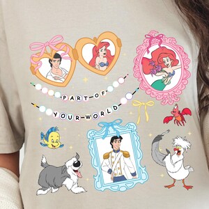 May include: Beige t-shirt with a vibrant graphic of Disney characters in decorative frames. The design includes Ariel, Prince Eric, and other characters, with the text "PART OF YOUR WORLD". A fun and whimsical design.