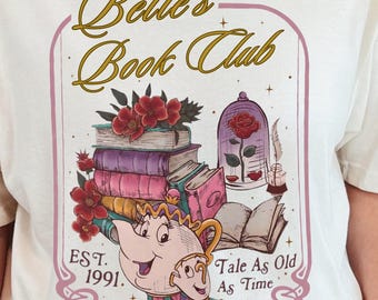Puede incluir: Camiseta color crema con un gráfico para el Club de Lectura de Bella. El diseño incluye una pila de libros, rosas, una rosa en una vitrina y una tetera. El texto incluye "Belle's Book Club", "EST. 1991" y "Tale As Old As Time".