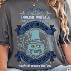 Peut inclure: T-shirt gris avec un motif graphique comportant le texte "WELCOME FOOLISH MORTALS" et "I AM YOUR HOST". Le motif comprend une figure fantomatique avec un chapeau haut de forme, dans des tons de bleu et de blanc.