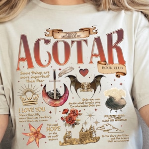 Pode incluir: Camiseta creme com o texto "ACOTAR BOOK CLUB" em fonte vermelho-alaranjada gradiente. A camiseta apresenta várias ilustrações, incluindo uma espada, asas e um castelo, com frases como "I LOVE YOU" e "HOPE".
