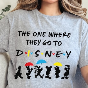May include: A light gray t-shirt with the text "THE ONE WHERE THEY GO TO DISNEY" in black. Below the text are silhouettes of Disney characters under colorful umbrellas. The shirt is a crew neck.