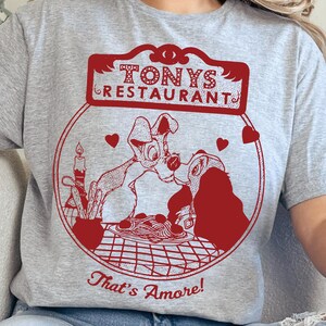 May include: A gray t-shirt featuring a red graphic of two dogs sharing spaghetti, with the text "Tony's Restaurant" above and "That's Amore!" below. The design is circular with hearts.