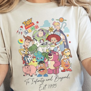 May include: Beige t-shirt featuring a colorful graphic of Toy Story characters, including Woody, Buzz Lightyear, and others. The text "Toy Story Land" is at the top, with "To Infinity and Beyond, Est. 1995" below. The shirt has short sleeves and a crew neck.