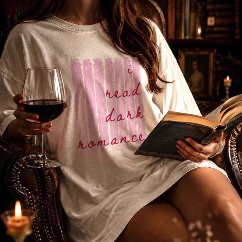 May include: Cream-colored long-sleeve shirt with the text "I read dark romance" in pink. The person is holding a glass of red wine and an open book. The scene is set in a dimly lit room with a bookshelf.