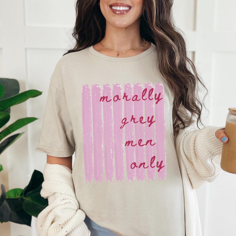 May include: A beige short-sleeved t-shirt featuring a graphic design of vertical light pink brushstrokes. Overlaid on the pink stripes is the dark pink cursive text that reads morally grey men only. The shirt has a crew neckline and a relaxed fit.
