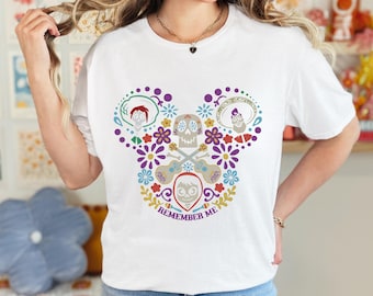 May include: White t-shirt featuring a colorful design in the shape of Mickey Mouse ears. The design includes skulls, guitars, and floral elements, with the words "Remember Me" at the bottom. The shirt is a casual wear item.