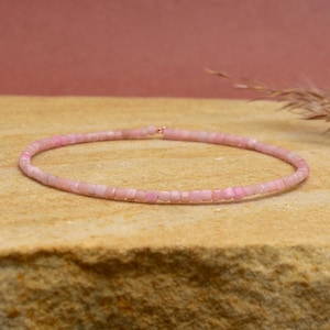 Puede incluir: Una delicada pulsera de cuentas rosas. La pulsera está compuesta por pequeñas cuentas cuadradas, ensartadas para formar una banda flexible. Las cuentas tienen un suave tono rosa pastel, y la pulsera se muestra sobre una superficie texturizada de color beige.