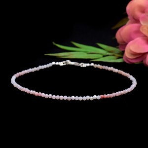 Könnte beinhalten: Zarter Armband aus kleinen, rosafarbenen Edelsteinperlen. Die Perlen sind mit einem silbernen Verschluss aufgefädelt. Das Armband hat eine weiche rosa Farbe, mit ein paar dunkleren rosa Perlen. Der Hintergrund ist schwarz.