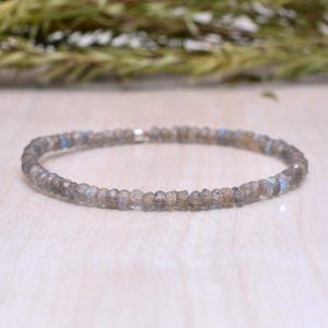May include: A delicate bracelet composed of small, faceted, gray-toned beads. The beads have subtle blue and iridescent flashes. The bracelet is strung on a thin, elastic band. The jewelry is displayed on a light-colored surface.