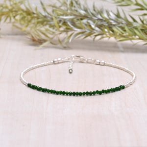 May include: A delicate silver anklet featuring a strand of small, vibrant green beads. The anklet has a simple design with a secure clasp. The background is a light wooden surface with greenery.