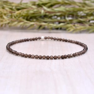 Pode incluir: Uma pulseira delicada composta por pequenas contas castanhas facetadas. As contas são enfiadas juntas, formando uma forma circular. A pulseira é exibida em uma superfície de madeira clara, com uma planta verde desfocada ao fundo.