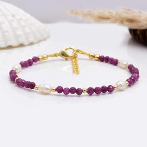 Könnte beinhalten: Ein zartes goldenes Armband mit einer Kette aus kleinen, facettierten, tiefrosa Edelsteinen, die mit cremeweißen Perlen und kleinen goldenen Perlen durchsetzt sind. Das Armband hat einen goldenen Verschluss.
