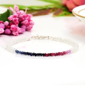 May include: A delicate bracelet featuring a gradient of colorful gemstones. The bracelet transitions from clear to blue, black, red, and pink beads. The clasp is silver. The bracelet is displayed on a white surface with pink flowers in the background.
