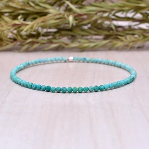Peut inclure: Un bracelet délicat en perles turquoises. Le bracelet est composé de petites perles rondes de couleur turquoise, enfilées sur un cordon fin et souple. Les perles ont un aspect légèrement facetté, et le bracelet est posé sur une surface en bois clair.