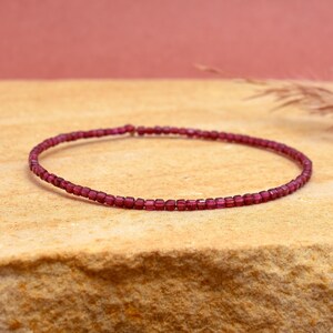 May include: A delicate, thin bracelet composed of small, rectangular, deep red beads. The bracelet is displayed on a textured, light brown surface, with a soft, reddish-brown background.
