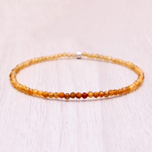 May include: A delicate bracelet with small, faceted beads in shades of yellow, orange, and red. The beads are strung together, with a silver clasp. The bracelet is displayed on a light-coloured wooden surface.
