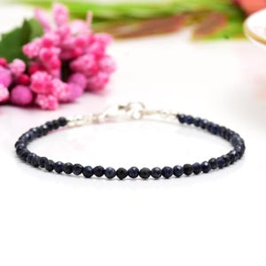 Könnte beinhalten: Ein zartes Armband mit kleinen, facettierten, dunkelblauen Perlen, die aufgefädelt sind. Das Armband hat einen silbernen Verschluss und wird vor einem weichen, weißen Hintergrund mit rosa Blumen im Hintergrund präsentiert.
