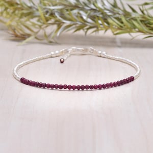May include: A delicate silver bracelet featuring a strand of small, faceted, deep red beads. The bracelet has a simple design with a secure clasp. The beads are evenly spaced along the silver chain, creating a subtle, elegant look.