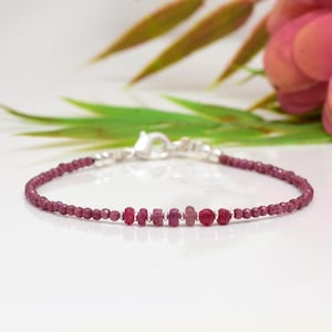 Puede incluir: Una pulsera delicada con pequeñas cuentas redondas de color rojo intenso y una serie de piedras más grandes, de forma irregular, de color rubí. La pulsera se asegura con un cierre plateado. El fondo incluye elementos florales verdes y rosas.