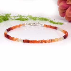 May include: A delicate bracelet featuring small, irregularly shaped gemstones in shades of red, orange, and white. The bracelet is strung on a silver chain and has a silver clasp. The gemstones are arranged in a gradient pattern, creating a visually appealing accessory.