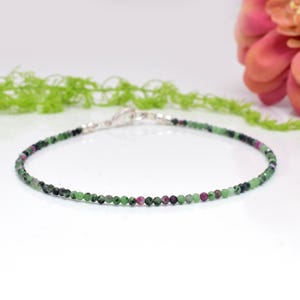 May include: A delicate bracelet made of small, round gemstones in green, black, and pink. The bracelet is strung on a thin, silver-coloured chain with a secure clasp. The jewellery is displayed on a white surface.