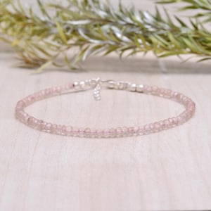 May include: A delicate bracelet featuring small, faceted pink gemstones strung together. The bracelet has a silver clasp and is displayed on a light-coloured wooden surface. The gemstones appear to be approximately 3mm in diameter.