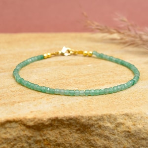 Puede incluir: Una delicada pulsera con pequeñas cuentas de color verde claro. La pulsera tiene detalles dorados en el cierre y los puntos de conexión. Las cuentas son de tamaño uniforme, creando un diseño simple y elegante. La pulsera se muestra sobre una superficie texturizada y beige.