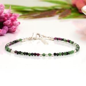 Może przedstawiać: Delikatna bransoletka z małymi, okrągłymi kamieniami szlachetnymi w odcieniach zieleni, czerni i bordowego. Bransoletka jest nawleczona na srebrny łańcuszek i ma srebrne zapięcie. Stylowa biżuteria.