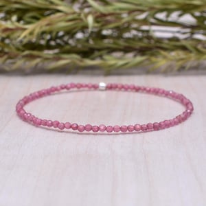 Op de afbeelding: Een delicate roze edelsteenarmband. De armband is gemaakt van kleine, ronde, roze kralen die aan elkaar zijn geregen met een zilveren sluiting. De armband wordt getoond op een licht houten oppervlak met groene bladeren op de achtergrond.