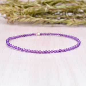 Könnte beinhalten: Eine zarte, lila Perlenkette. Die Kette besteht aus kleinen, runden, facettierten Perlen, die zu einem vollständigen Kreis aufgereiht sind. Die Perlen sind in einem leuchtenden Lilaton gehalten, und die Kette wird auf einer hellen Oberfläche präsentiert.