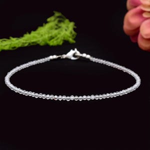 Pode incluir: Uma pulseira delicada com pequenas contas claras e facetadas. A pulseira tem um fecho prateado e é exibida sobre um fundo preto com detalhes verdes e rosa. Comprimento aproximado: 18 cm.