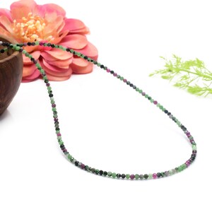 May include: A delicate necklace composed of small, round beads in shades of green, black, and pink. The necklace is displayed against a white background, with a pink and orange flower and green foliage in the background. The necklace is a jewelry item.