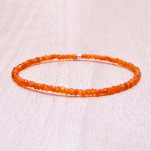 Könnte beinhalten: Ein zartes Armband aus kleinen, orangefarbenen Edelsteinperlen. Die Perlen sind aufgefädelt und bilden eine kreisförmige Form. Ein kleiner, silberfarbener Verschluss sichert das Armband. Das Armband wird auf einer hellen Holzoberfläche präsentiert.
