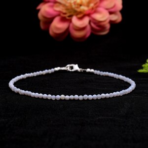 Op de afbeelding: Een delicate, lichtblauwe armband met een zilveren sluiting. De armband bestaat uit kleine, ronde kralen en wordt gepresenteerd op een zwart oppervlak. Op de achtergrond een roze bloem.
